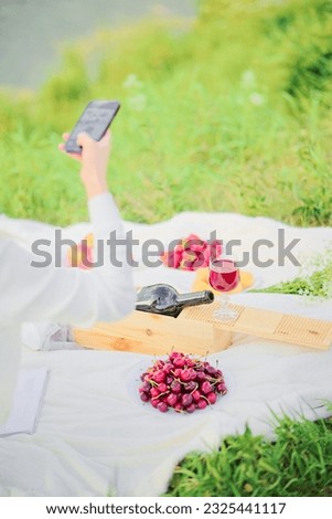 Woman taking pictures of food, wine, cherries, bottle, fruit on her smartphone at a picnic.