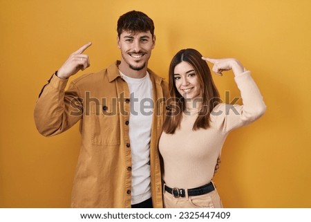 Young hispanic couple standing over yellow background smiling pointing to head with both hands finger, great idea or thought, good memory 
