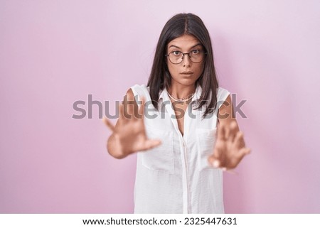 Brunette young woman standing over pink background wearing glasses afraid and terrified with fear expression stop gesture with hands, shouting in shock. panic concept.  Royalty-Free Stock Photo #2325447631