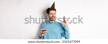 Celebration and holidays concept. Cheerful young man making wish on birthday, holding fingers crossed and wearing party hat, standing with bday cake, white background.