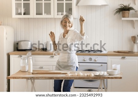 Cheerful active pretty mature woman dancing at floury kitchen table with bakery food products, preparing fresh homemade dessert, enjoying baking, domestic culinary hobby, having fun