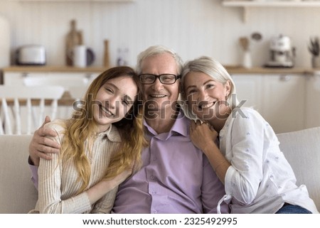 Cheerful older grandfather enjoying leisure with wife and grandkid, looking at camera, smiling, laughing, hugging family in sofa. Home portrait of happy grandparents and teenage kid