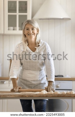 Happy pretty blonde mature baker woman in apron rolling raw dough on wooden rustic kitchen table, baking fresh homemade buns, looking at camera, smiling. Vertical shot portrait Royalty-Free Stock Photo #2325493005