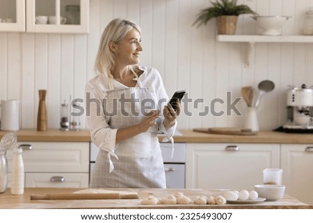 Happy pretty blonde mature housewife in apron browsing cooking recipe on smartphone, preparing homemade bakery food, holding mobile phone, looking away, smiling, thinking