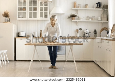 Mature blonde domestic baker woman cooking at home, rolling raw dough, standing at floury table in traditional white kitchen interior, baking fresh homemade pastry meal for breakfast Royalty-Free Stock Photo #2325493011