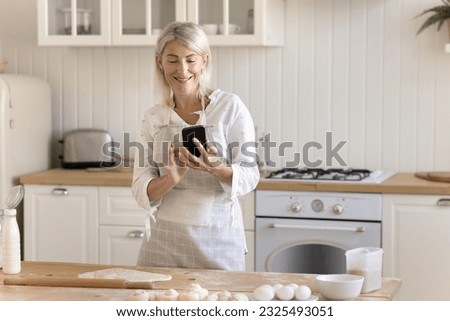 Cheerful baker blogger woman taking picture of baking process on smartphone for cooking blog, holding mobile phone over floury kitchen table with raw dough, roller, using Internet technology