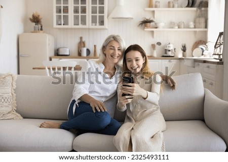 Cheerful mature granny and young teenage kid girl using media application on smartphone together, enjoying Internet technology, family communication, talking selfie photo
