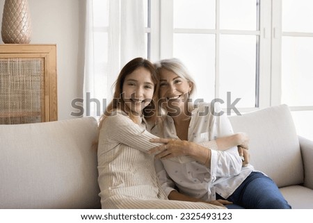 Beautiful grandma and teenager granddaughter posing at home, sitting on couch together, hugging, enjoying shooting, family leisure, hugging, looking at camera, smiling, laughing