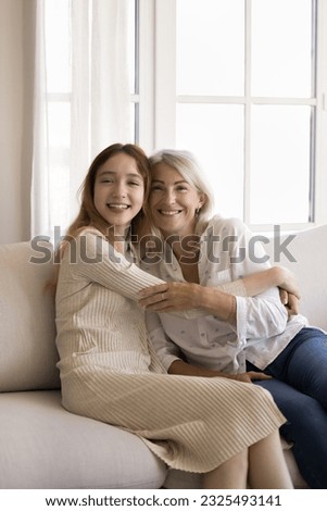 Happy pretty teenager girl embracing cheerful blonde grandmother on home sofa, looking at camera, smiling, laughing, posing for family portrait. Granddaughter and grandma vertical shot