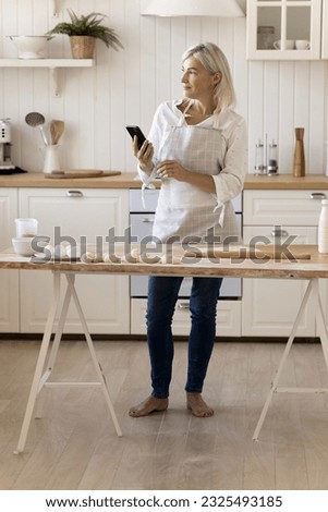 Thoughtful mature blonde baker woman using mobile phone at kitchen table with bakery ingredients, baking homemade buns, holding smartphone, looking away, thinking. Vertical shot