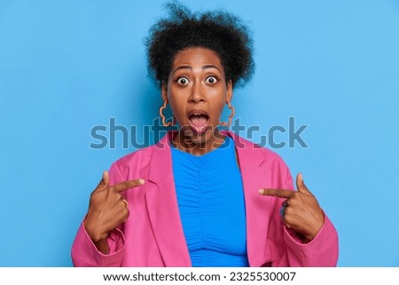 Young African-American woman in blue and pink clothes stands against blue wall, pointing index fingers on herself, lucky time concept, copy space