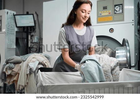 Worker of laundry shop loading industrial washing machine with clothes Royalty-Free Stock Photo #2325542999