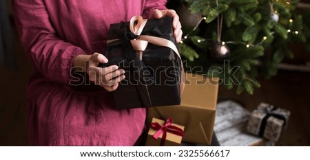 A woman in a burgundy dress holds a large gift with a bow against the background of a Christmas tree. Gifts for the New Year with a place for text