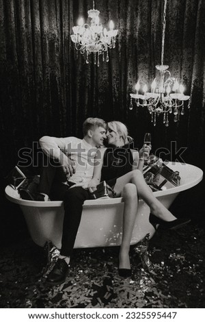 A romantic woman is lying in the bathroom with New Year's gifts, her husband is standing next to her with glasses of champagne. Merry Christmas. Black and white photo with artistic noises