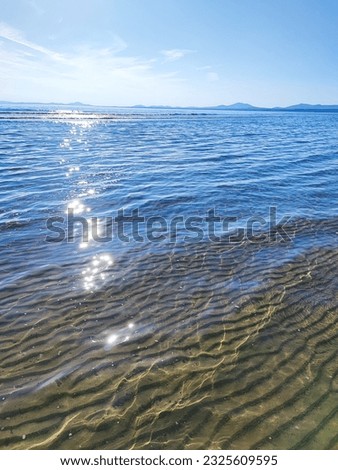 Sun reflecting on waves with blue silhouettes of mountains in the background and underwater ripples in the sand in the foreground.