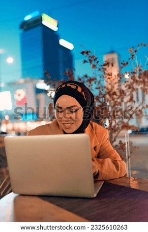In the enchanting ambiance of a nocturnal cityscape, a hijab-clad girl engrossed in her laptop creates a mesmerizing image, embodying the fusion of technology, empowerment, and the vibrant urban