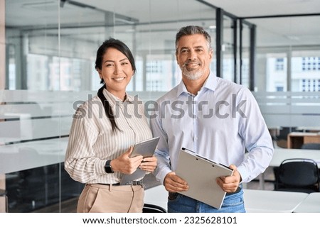 Happy confident professional mature Latin business man and Asian business woman corporate leaders managers standing in office, two diverse colleagues executives team posing together, portrait. Royalty-Free Stock Photo #2325628101