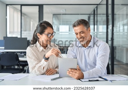 Two diverse business team people talking and working in office on corporate project at meeting. Young Asian employee and mid aged Latin executive looking at digital tablet company technology device.