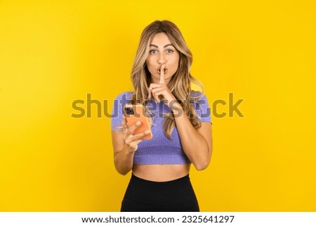 Young beautiful blonde woman wearing sportswear over yellow studio background  holding modern gadget ask not tell secrets Royalty-Free Stock Photo #2325641297