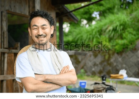 A Japanese farmer stands in the yard of an old house in the Japanese countryside with his arms folded and a smile on his face. The photo shows the upper half of his body, looking at the camera. Royalty-Free Stock Photo #2325695341