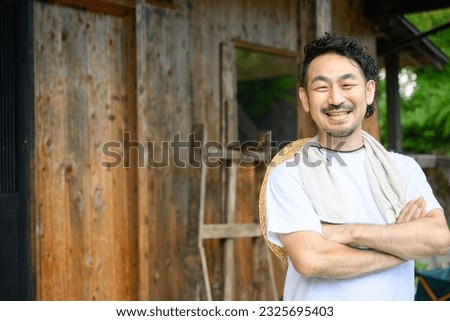 A Japanese farmer stands in the yard of an old house in the Japanese countryside with his arms folded and a smile on his face. The photo shows the upper half of his body, looking at the camera. Royalty-Free Stock Photo #2325695403