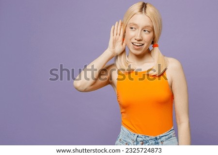 Young curious nosy blonde woman wear orange tank shirt casual clothes try to hear you overhear listening intently isolated on plain pastel light purple background studio portrait. Lifestyle concept