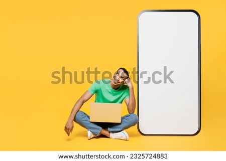 Full body young IT man he wears casual clothes green t-shirt hat big huge blank screen mobile cell phone smartphone with area hold use work on laptop pc computer isolated on plain yellow background