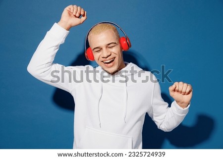 Young vivid happy cheerful dyed blond man of African American ethnicity wear white hoody headphones listen music dance raise up hands isolated on plain dark royal navy blue background studio portrait