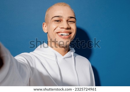 Close up smiling fun young dyed blond man of African American ethnicity wear white hoody doing selfie shot pov on mobile cell phone isolated on plain dark royal navy blue background studio portrait