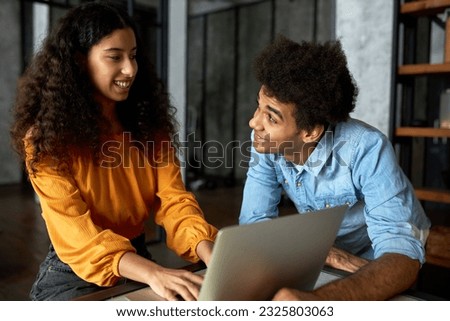 Indoor image of happy african american co-workers talking, flirting looking at each other smiling while girl working on laptop, guy with afro hair in denim shirt joking, making her laugh