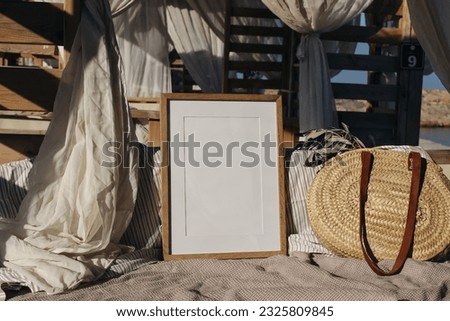 Blank picture frame, poster mockup in beach lounger, gazebo with canopy in resort at sea. Wicker straw bag with green olive tree branches in sunlight. Summer vacation, traveling concept.