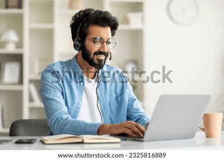 Hotline Operator. Portrait Of Indian Customer Support Manager Man In Headset Working With Laptop At Home Office, Smiling Eastern Male Consulting Clients, Sitting At Desk With Computer, Free Space Royalty-Free Stock Photo #2325816889
