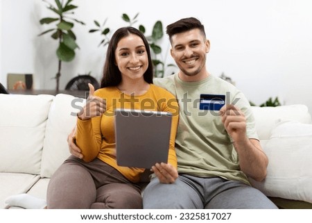 Happy smiling beautiful young couple paying for goods and services on Internet from home, sitting on couch in living room, using modern digital pad and bank card, copy space. E-commerce, retail