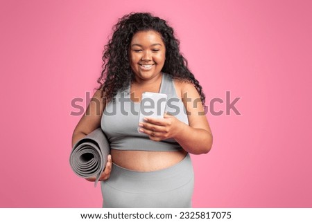 Smiling black chubby woman in sportswear with mat chatting on smartphone, standing on pink studio background. Sports app, blog for weight loss and body care, trainer lesson remotely