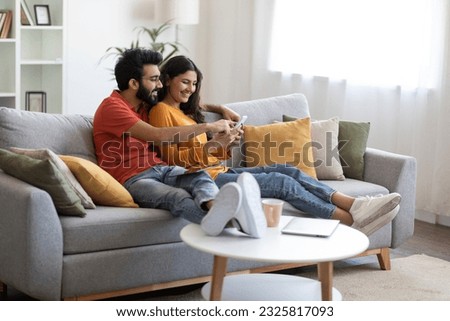 Beautiful Young Indian Couple Using Mobile Phone And Embracing Together At Home, Happy Eastern Spouses Relaxing On Couch With Modern Smartphone, Browsing New App Or Shopping Online, Copy Space Royalty-Free Stock Photo #2325817093