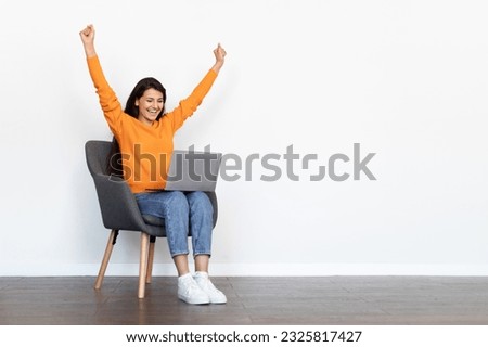Thrilled emotional beautiful millennial hindu woman wearing casual outfit sitting in armchair over white blank wall, looking at computer laptop screen, gesturing raising hands up, copy space Royalty-Free Stock Photo #2325817427