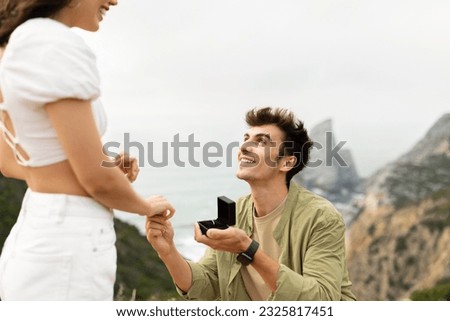 Engagement proposal. Happy young man with ring making marriage offer with beautiful coastline view, standing on his knee on the cliff. She said yes on the top Royalty-Free Stock Photo #2325817451
