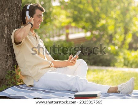 Positive relaxed happy handsome millennial man wearing casual outfit chilling on blanket under tree at park, enjoying sunny summer day, using cell phone and wireless headphones, copy space