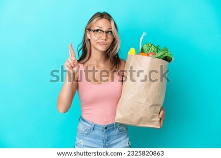 Young Russian woman holding a grocery shopping bag isolated on blue background with fingers crossing and wishing the best