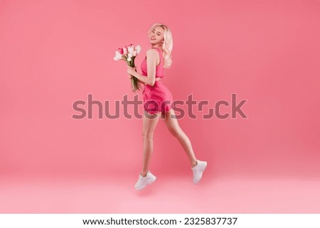 Full body length shot of happy young lady jumping holding bouquet of flowers and smiling at camera, celebrating birthday, being in good mood over pink studio background, free space