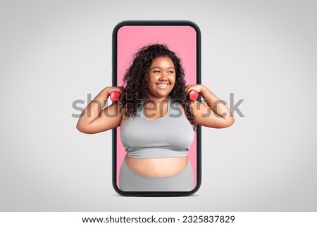 Positive black oversize lady in sportswear doing exercises with hand dumbbells on big phone screen, grey studio background, banner. Sports app, blog for weight loss and body care