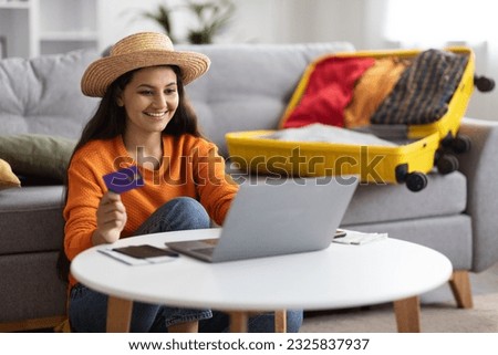 Cheerful excited beautiful millennial hindu woman in casual outfit and wicker hat sitting on floor, holding bank card, booking hotel or plane ticket online, using laptop at home, copy space Royalty-Free Stock Photo #2325837937