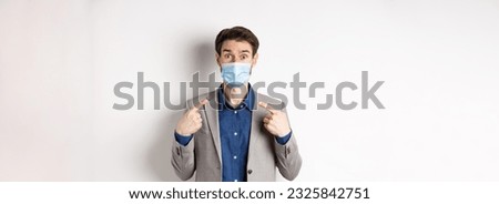 Covid-19, pandemic and business concept. Excited businessman in suit pointing at his medical mask and looking at camera, white background.