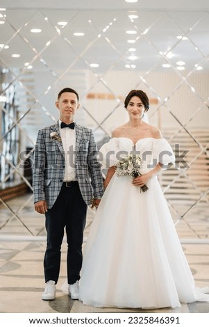 Beautiful bride in wedding dress with bouquet and groom posing near mirror. Full length of attractive young woman wearing dress and man while ceremony. Big mirror wall. Marriage.