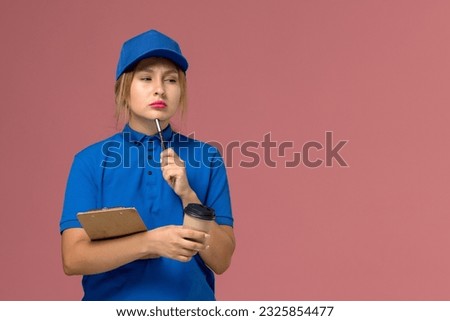 front view female courier in blue uniform posing holding cup of coffee and notepad thinking on pink desk service uniform delivery girl