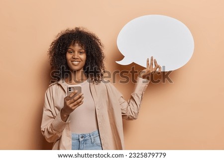 Cheerful African woman dressed in casual attire holds smartphone amidst communication bubble smiles pleasantly wears shirt and jeans isolated over brown background. People and advertisement.