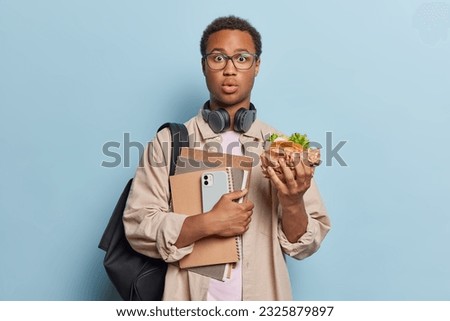 Waist up shot of shocked youngster eats sandwich during break at school carries notebooks smartphone and backpack reacts to something very surprising stands against blue background. Fast food Royalty-Free Stock Photo #2325879897