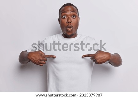 Photo of shocked dark skinned man points both index fingers at empty space of t shirt stares with disbelief has bugged eyes holds breath isolated over white background. Place your logo here.