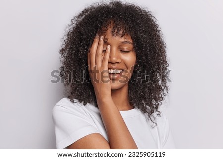 Curly haired woman radiates positivity as she covers half her face with her palm eyes closed in blissful contentment dressed in casual tshirt isolated on white background. People and happiness concept Royalty-Free Stock Photo #2325905119