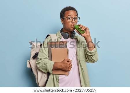 Photo of surprised dark skinned man drinks fresh vegetable smoothie from bottle carries notepads poses with rucksack wears spectacles and shirt isolated over blue background being on way to college Royalty-Free Stock Photo #2325905259
